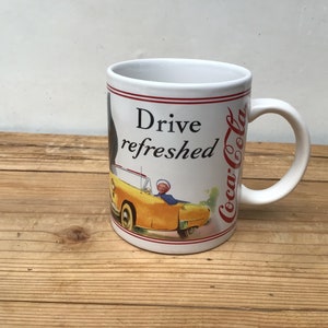 May include: White ceramic mug with a red and white Coca-Cola logo and the text "Drive refreshed". The mug features a vintage illustration of a yellow car with a person driving.