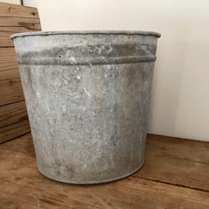May include: A weathered, galvanized metal bucket with a cylindrical shape. The bucket has a rolled rim and a slightly tapered body. The metal has a mottled gray appearance with signs of age and use. The bucket is sitting on a wooden surface.