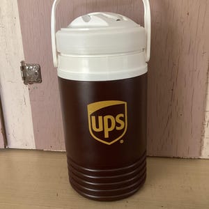 May include: A brown and white insulated beverage cooler with a white handle. The cooler has the UPS logo in gold on the front. The cooler is cylindrical with a ridged base.
