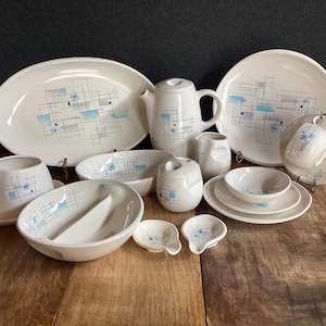 May include: A collection of off-white ceramic dinnerware featuring a blue and grey geometric grid pattern with starburst accents. The set includes a large oval platter, a round plate, a teapot, a creamer, a sugar bowl, bowls, and small dishes.