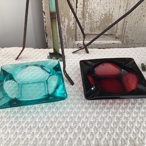 May include: Two square glass ashtrays, one in a translucent light turquoise blue and the other in a deep translucent red. Each ashtray features four rounded indentations along the edges, designed to hold items securely in place.