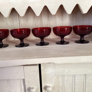 May include: Five vintage ruby red glass dessert bowls with black stems are displayed on a white shelf. The bowls have a rounded shape and are evenly spaced. The background features a white cabinet with a decorative top.