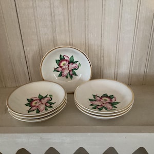 May include: A set of vintage white ceramic bowls with a gold rim. Each bowl features a pink and green floral design in the center. The bowls are arranged in stacks of three and four, with one bowl standing alone.
