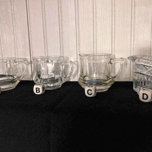 May include: Four clear glass creamer pitchers with different designs. The pitchers are arranged in a row from left to right. The pitcher on the left is plain, the second pitcher has a textured design, the third pitcher has a simple rim, and the fourth pitcher has a more intricate design.