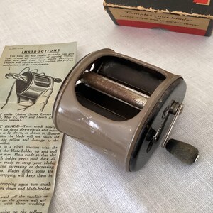 May include: Vintage metal razor blade sharpener with a crank handle and wooden grip. The device is a light brown color with a black base and a box that says "Twinplex your blades". Instructions are visible.