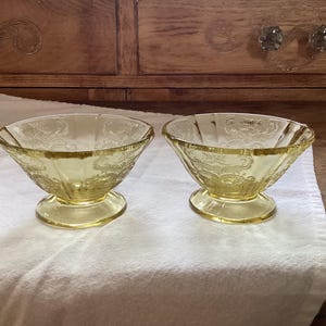May include: Two vintage yellow glass dessert bowls with a pedestal base. The bowls have a decorative floral pattern and are displayed on a white cloth. The background features a wooden cabinet.