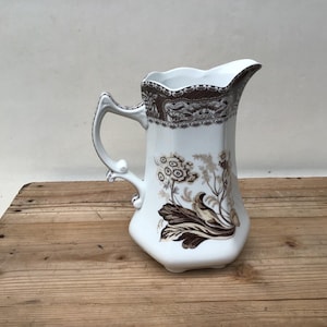 May include: White ceramic pitcher with a brown and gold floral design. The pitcher has a scalloped rim and a handle.