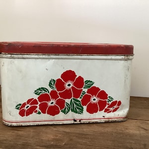 May include: A rectangular, vintage metal storage tin with a red lid and a white body. The white body features a floral design of red flowers with green leaves. The tin sits on a wooden surface.