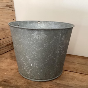 May include: A galvanized metal bucket with a slightly tapered shape. The bucket has a rolled rim and a small hole near the top edge. It sits on a wooden surface, with a wooden wall in the background.