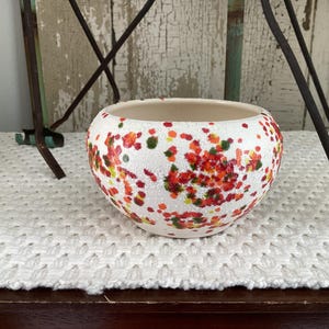 May include: A round ceramic bowl with a white base featuring a textured, speckled pattern of red, orange, and green paint splatters. The interior of the bowl is smooth and off-white, while the exterior surface has a granular, matte finish.