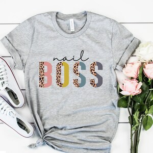 May include: Gray t-shirt with the text "nail BOSS" in colorful letters with a leopard print pattern. The letters are in different colors, including pink, yellow, blue, and gray.