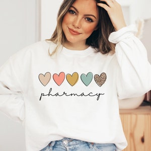 May include: White sweatshirt with a row of five colorful hearts and the word "pharmacy" written below. The hearts are pink, peach, yellow, teal, and brown with a leopard print.