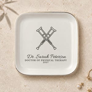 May include: A square white ceramic trinket dish with a metallic gold rim. The center features a black line drawing of two crossed crutches above the text Dr. Sarah Peterson, DOCTOR OF PHYSICAL THERAPY, and the year 2027 printed in black ink.