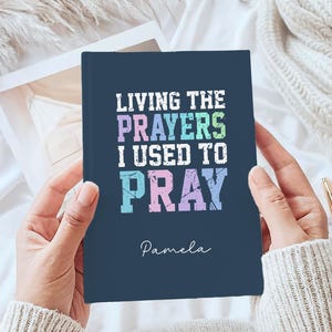 May include: A navy blue journal with the text "LIVING THE PRAYERS I USED TO PRAY" in colourful letters. The name "Pamela" is written below the text. The journal is held in hands.
