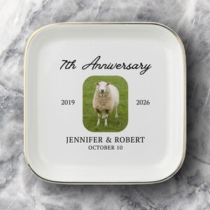 May include: A square white ceramic trinket dish with a gold-toned rim. The center features a photograph of a sheep in a green field, surrounded by the text 7th Anniversary, 2019, 2026, JENNIFER & ROBERT, and OCTOBER 10 in black lettering.