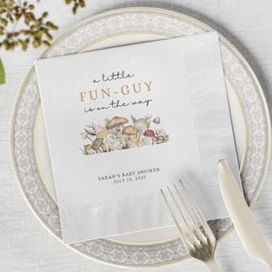 May include: White paper napkin with the text "a little FUN-GUY is on the way" in gold and black. A watercolor illustration of mushrooms and plants is below the text. The napkin is on a plate with a fork and knife.