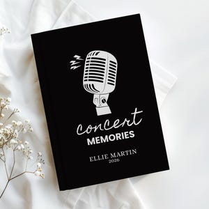 May include: A black journal featuring a white microphone illustration and the text "Concert Memories." The journal also displays the name "Ellie Martin" and the year "2026." It rests on a white surface, accompanied by white flowers.