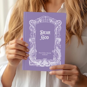 May include: A lavender journal with the words "DEAR GOD" in elegant script, framed by ornate white scrollwork. The text "Prayers from Sarah" is below. The journal is held in front of a white shirt.