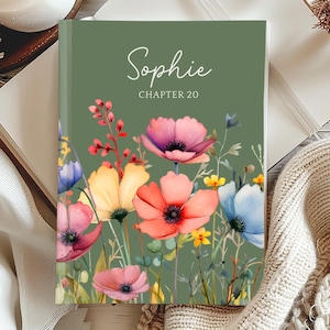 May include: A hardcover book with a green cover featuring the name "Sophie" and "Chapter 20" in white script. The cover is adorned with watercolor flowers in shades of pink, orange, yellow, and blue. The book is open on a wooden surface.