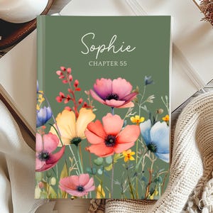 May include: A hardcover journal with a green cover featuring the name "Sophie" and "Chapter 55" in white script. The cover is adorned with a watercolor floral design in shades of pink, orange, yellow, and blue. The journal is open on a wooden surface.