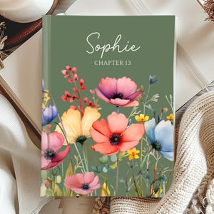 May include: A hardcover book with a green cover featuring the name "Sophie" and "Chapter 13" in white script. The cover is adorned with a watercolor floral design in shades of pink, orange, yellow, and blue. The book is open on a wooden surface.