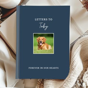 May include: A dark blue hardcover journal with the text "LETTERS TO Toby" printed in white and a square photograph of a golden-colored dog lying in green grass. The bottom of the cover features the text "FOREVER IN OUR HEARTS" in white.