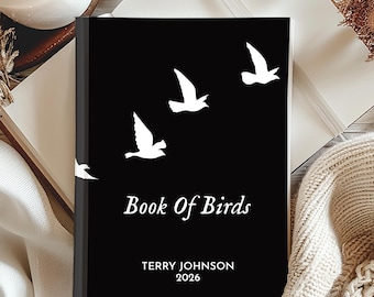 Puede incluir: Un libro negro titulado "Book Of Birds" con siluetas de pájaros blancos. La portada del libro también incluye el nombre del autor, Terry Johnson, y el año 2026. El libro está sobre una superficie blanca con una vela y otros elementos decorativos.