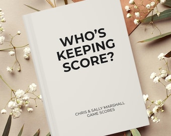 May include: A white book with the title "Who's Keeping Score?" and the text "Chris & Sally Marshall Game Scores" on the cover. The book is lying on a beige surface with white flowers and green leaves.