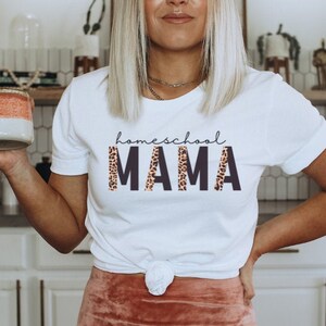 Op de afbeelding: Wit T-shirt met de tekst "homeschool MAMA" in zwarte letters met een luipaardprint.