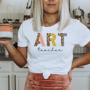 K&ouml;nnte beinhalten: Wei&szlig;es T-Shirt mit dem Wort "ART" in Leopard-Print, rosa, gelben und grauen Buchstaben. Der Text "teacher" ist darunter in Kursivschrift geschrieben.