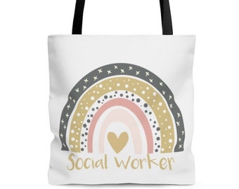 May include: A white tote bag with black handles features a rainbow design in shades of pink, gray, and gold. The rainbow has a small heart in the center. The text "Social Worker" is printed in gold below the rainbow.