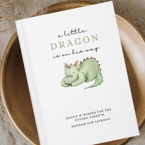May include: A white baby shower advice book with the text "a little DRAGON is on his way" and a watercolor illustration of a green dragon. The book includes the text "ADVICE & WISHES FOR THE FUTURE PARENTS HANNAH AND JACKSON".
