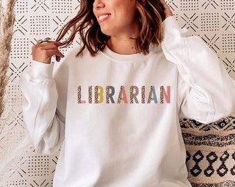 May include: White sweatshirt with the word "LIBRARIAN" printed in colorful letters with a leopard print pattern. The sweatshirt is being worn by a person sitting on a white couch.