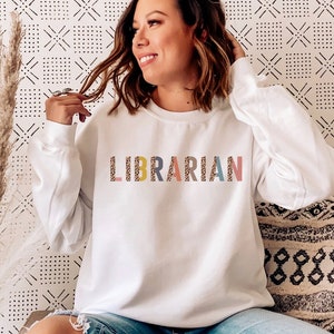Peut inclure: Sweat-shirt blanc avec le mot "LIBRARIAN" imprim&eacute; en lettres color&eacute;es avec un motif l&eacute;opard. Le sweat-shirt est port&eacute; par une personne assise sur un canap&eacute; blanc.