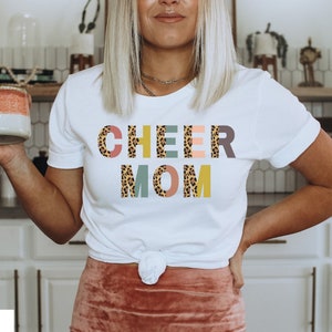 以下が含まれることがあります： レオパード柄のカラフルな文字で「CHEER MOM」と書かれた白いTシャツです。