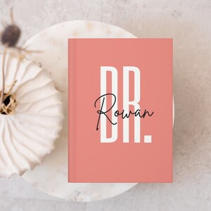 May include: A coral-colored journal with the text "DR. Rowan" in white and black. The journal is on a white marble-like surface, with a decorative white ceramic piece and dried plant in the background. The journal is a notebook or diary.