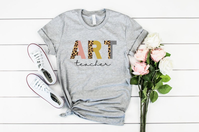 K&ouml;nnte beinhalten: Ein graues T-Shirt mit dem Wort "ART" in rosa, gelben und braunen Leopardenmuster-Buchstaben. Das Wort "teacher" ist in schwarzer Schrift unter den Buchstaben geschrieben.