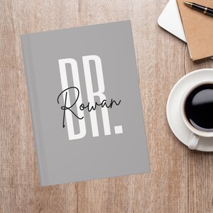 May include: A gray journal with the text "DR. Rowan" in white and black. The journal is on a wooden surface next to a cup of coffee and a notebook.