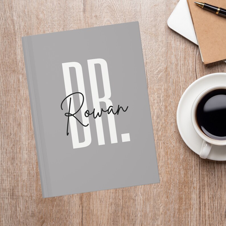May include: A gray journal with the text "DR. Rowan" in white and black. The journal is on a wooden surface next to a cup of coffee and a notebook.