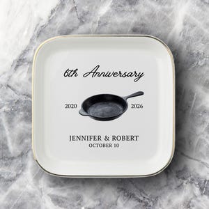 May include: A square white ceramic trinket dish with a gold-toned rim. The center features a black cast iron skillet graphic, the text 6th Anniversary, the years 2020 and 2026, the names JENNIFER & ROBERT, and the date OCTOBER 10 in black lettering.