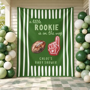 May include: A green fabric backdrop featuring white vertical stripes, a brown football, and a red foam finger graphic. The text reads: a little ROOKIE is on the way, CHLOE'S BABY SHOWER. The banner is framed by clusters of green and white balloons.