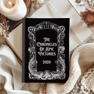 Puede incluir: Un diario negro con el título "The Chronicles of Epic Victories 2026" en escritura blanca. La portada presenta un diseño de marco decorativo y ornamentado. El diario está sobre una superficie blanca con una vela y otros elementos decorativos.