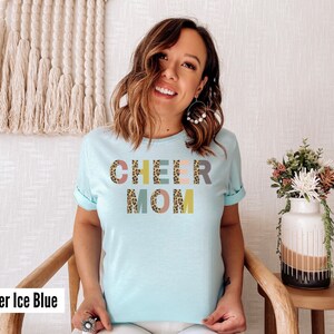 以下が含まれることがあります： レオパード柄のカラフルな文字で「CHEER MOM」と書かれた薄いブルーのTシャツ。