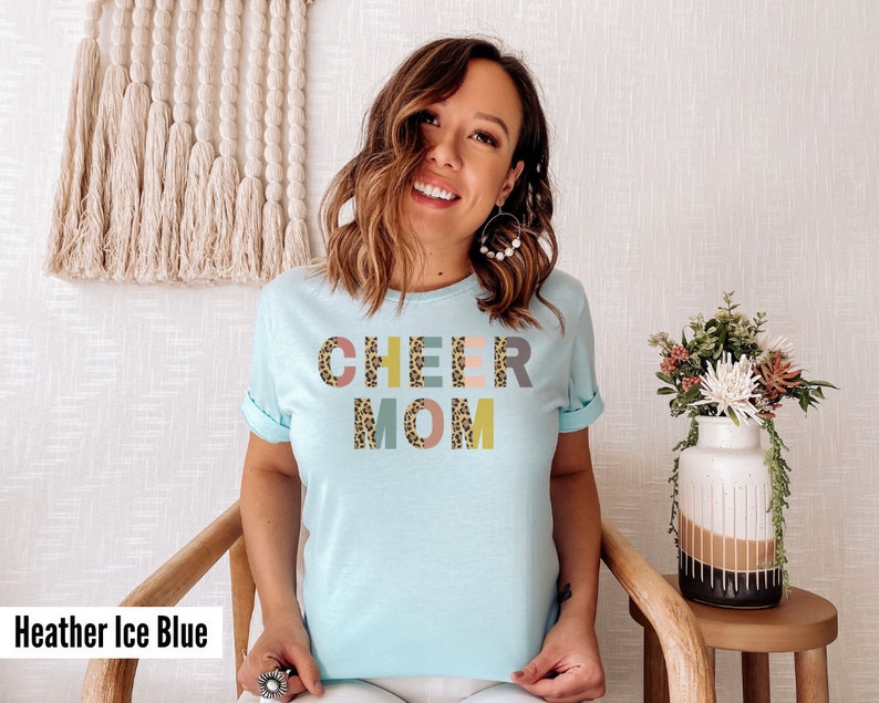 以下が含まれることがあります： レオパード柄のカラフルな文字で「CHEER MOM」と書かれた薄いブルーのTシャツ。