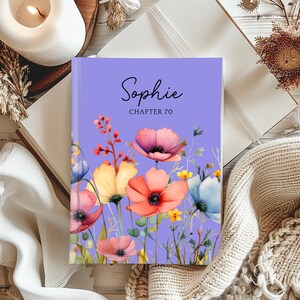 May include: A lilac journal with the name "Sophie" and "CHAPTER 70" in black script. The cover displays watercolour flowers in pink, orange, yellow, and blue. The journal is open on a white surface.