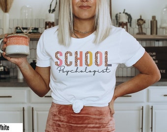 May include: White t-shirt with the text "SCHOOL Psychologist" in pink, brown, and yellow letters with a leopard print pattern. The letters are stacked on top of each other.
