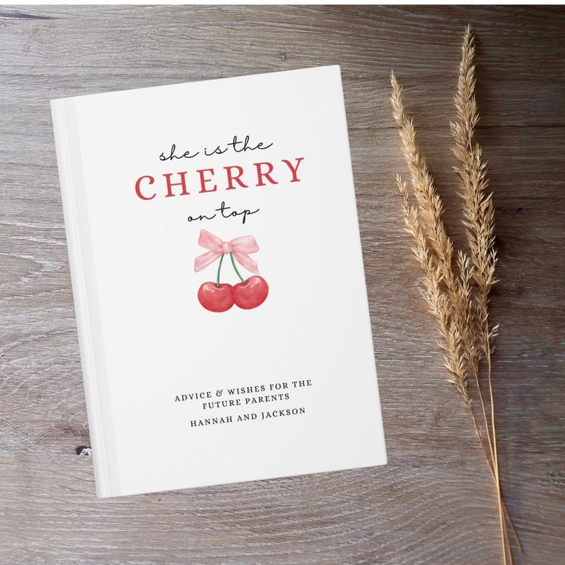May include: White book with the text "she is the CHERRY on top" in black and red lettering. A pink ribbon with two red cherries are illustrated on the cover. The text "ADVICE & WISHES FOR THE FUTURE PARENTS HANNAH AND JACKSON" is printed in black at the bottom of the cover.