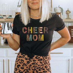 以下が含まれることがあります： レオパード柄のカラフルな文字で「CHEER MOM」と書かれた黒いTシャツ。文字はそれぞれ異なる色で、文字の背景はレオパード柄です。