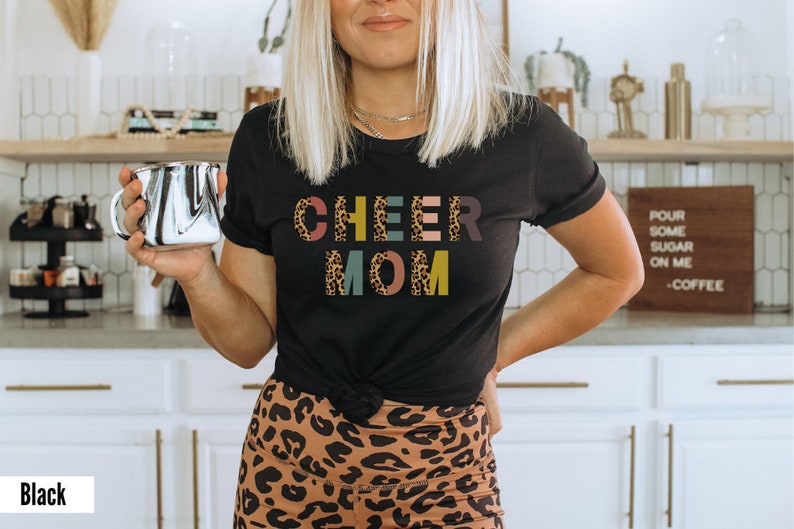 以下が含まれることがあります： レオパード柄のカラフルな文字で「CHEER MOM」と書かれた黒いTシャツ。文字はそれぞれ異なる色で、文字の背景はレオパード柄です。