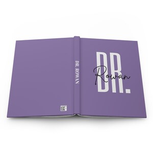May include: A lavender journal with the text "DR. Rowan" in white block letters and a handwritten-style signature. The spine reads "DR. ROWAN" vertically. A small barcode is visible on the lower left corner.